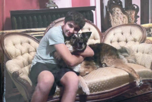 Photograph of Rocky Delfs sitting on a couch, hugging a dog and smiling into the camera.