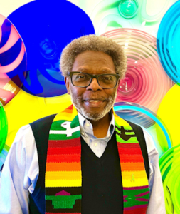 Rev. Isaac Ruffin, an African American male, wearing a colorful scarf representing Black heritage, standing in front of an art display of glass plates in various shades of primary colors.