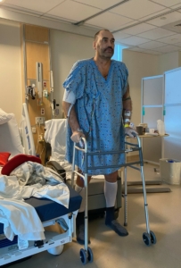 A tall man in a hospital room, wearing a hospital gown and standing with a walker.