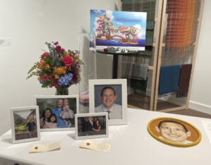 A display table with framed portraits, a vase filled with flowers and a rendering of the OneLegacy Donate Life Rose Parade float 2025.