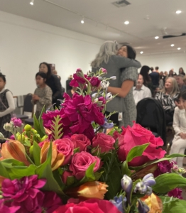 A bouquet of flowers in the foreground and a woman getting a hug in the background.